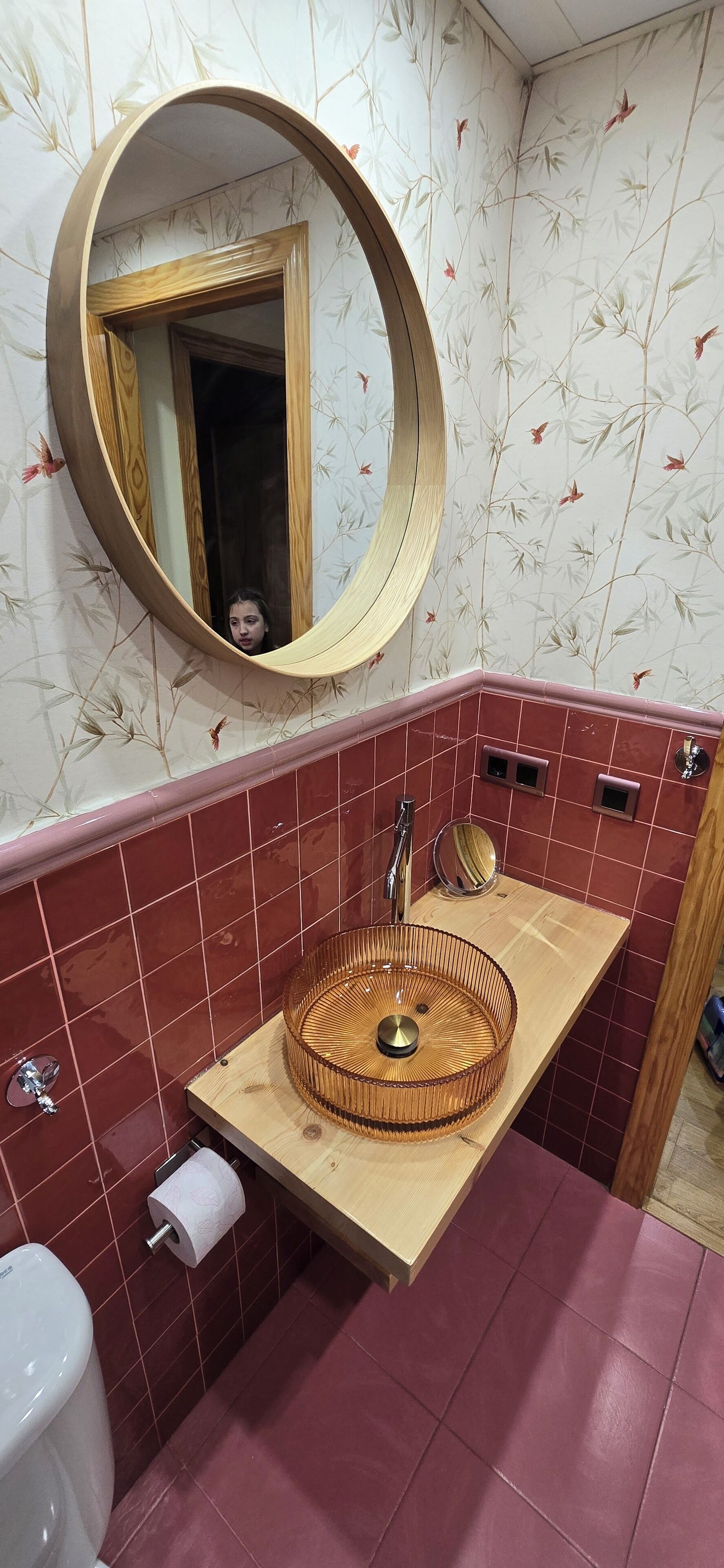 A bathroom with a round mirror, a wooden vanity, a sink with a circular basin, and red-tiled walls.