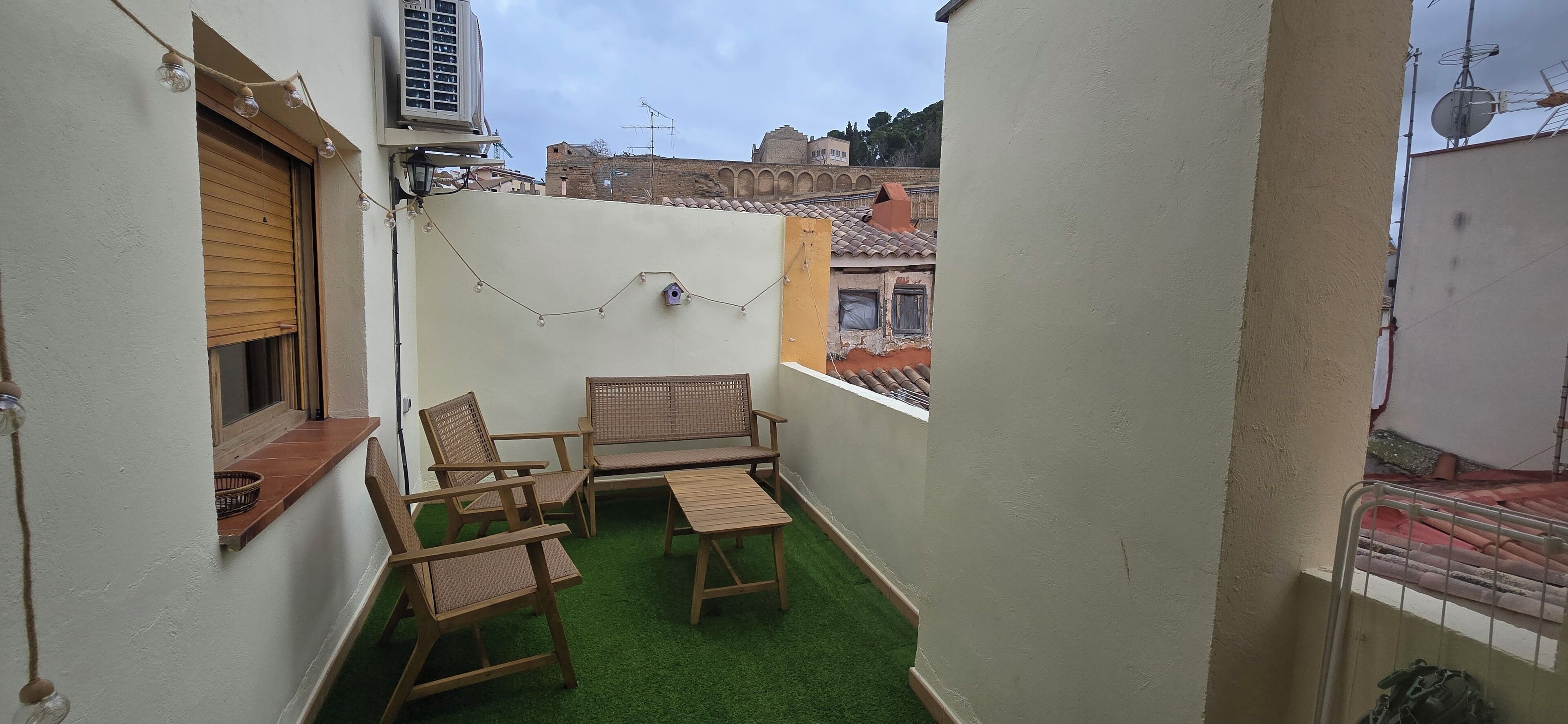 A small balcony with a green artificial turf, wooden furniture, and a string of lights.