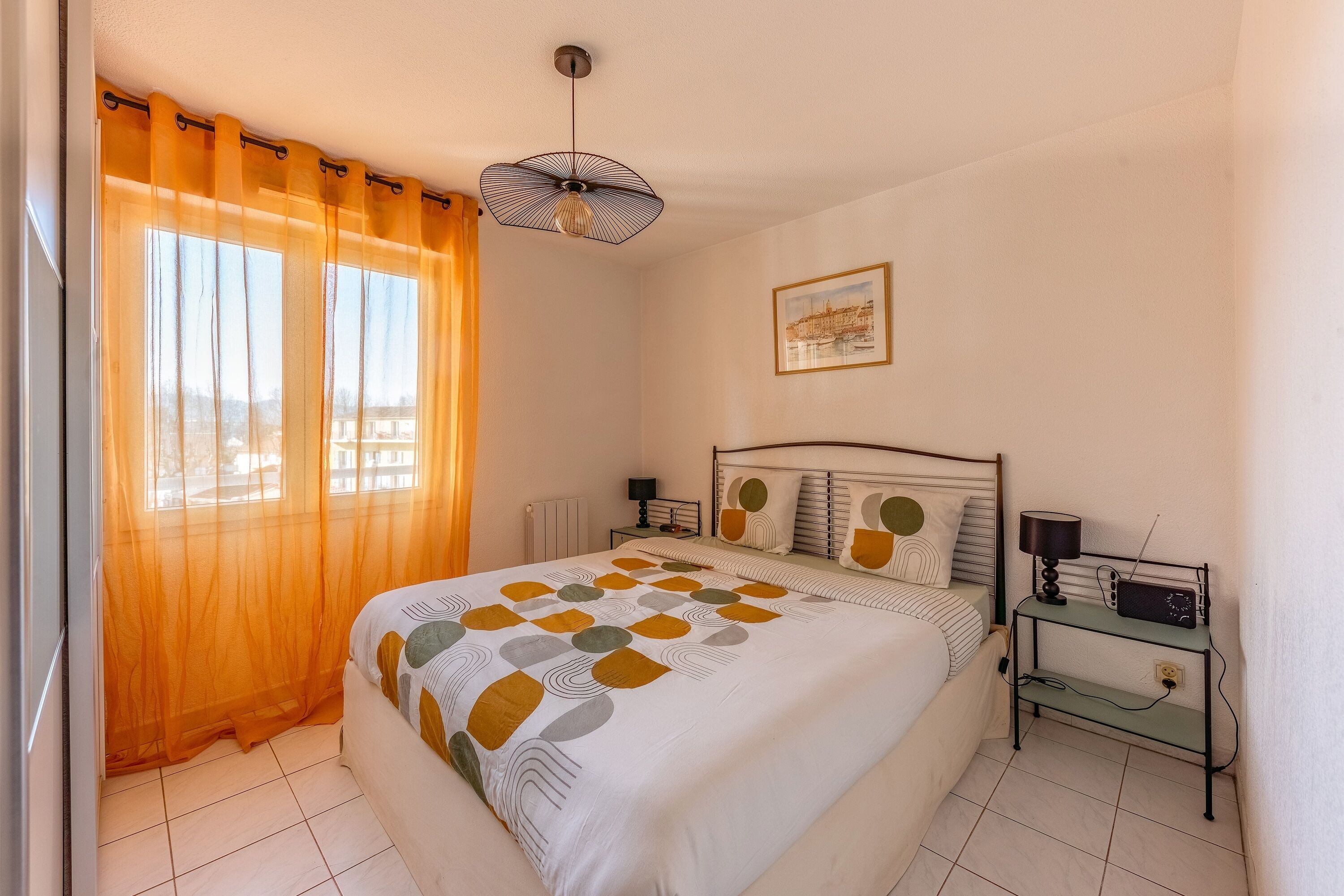 A bedroom with a bed, a nightstand, a lamp, a radio, and a window with sheer curtains.