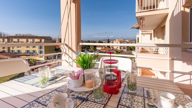 Ein Balkon mit Tisch für zwei Personen, ausgestattet mit einem Teekessel, Tassen und einer roten Kaffeemaschine. Der Balkon bietet Blick auf eine Stadtlandschaft mit Gebäuden und blauem Himmel.