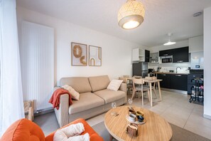 Ein modernes Wohnzimmer mit Sofa, Couchtisch und Essbereich. Im Hintergrund ist eine Küche mit Geräten und Schränken zu sehen.