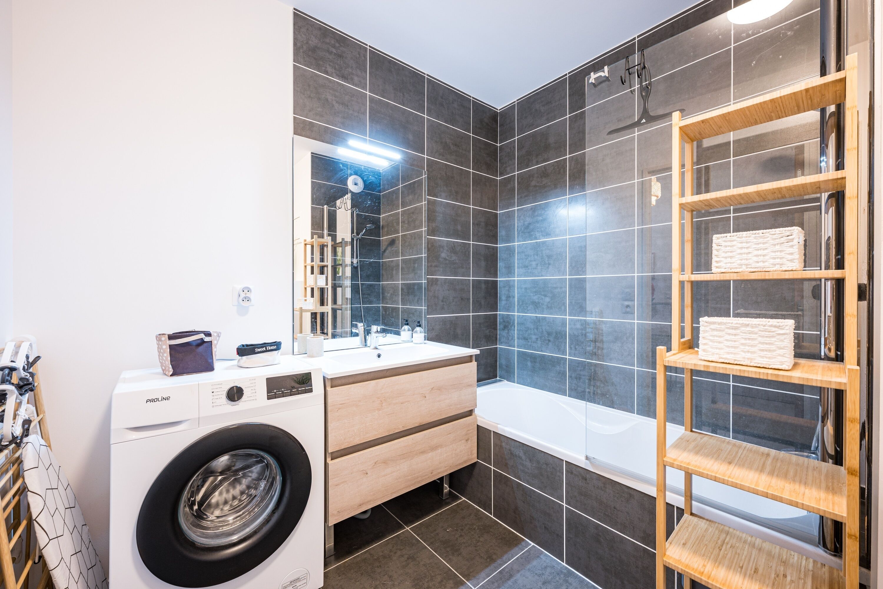 A modern bathroom with a washing machine, a bathtub, a wooden shelf, and a large mirror.