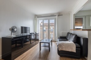 A modern living room with a black leather sofa, a glass coffee table, a black TV stand with a flat-screen TV, a standing fan, and a large window with a view of buildings. - Le Havre Aubervilliers - A 2 pas du métro (Aubervilliers)