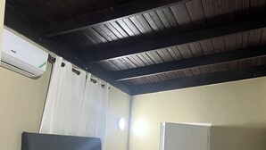 Un lit simple avec une literie blanche, une tête de lit sombre, un rideau blanc, une porte blanche et un plafond avec des poutres en bois.
