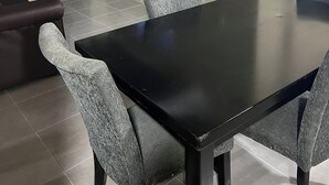 Un coin repas avec une table noire et quatre chaises grises, un téléviseur fixé au mur et un rideau foncé.