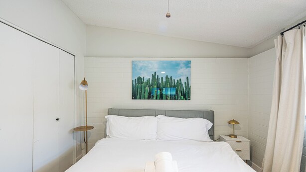 A neatly made bed with white linens and a rolled towel, a bedside table with a lamp, and a painting of cacti on the wall. - Dwell (Scottsdale)