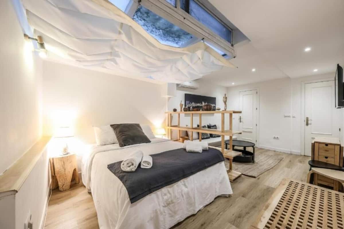 A modern bedroom with a skylight, a bed with a dark headboard, a wooden shelf, and a small table.
