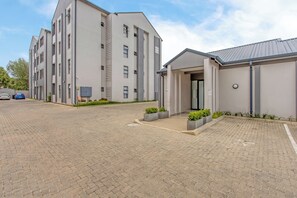 A multi-story building with a modern design, featuring a paved driveway and small landscaped areas with plants. - Wifi, Washer, Smart TV, Work station, Balcony, Pool, Free parking, 24hr security (Midrand)