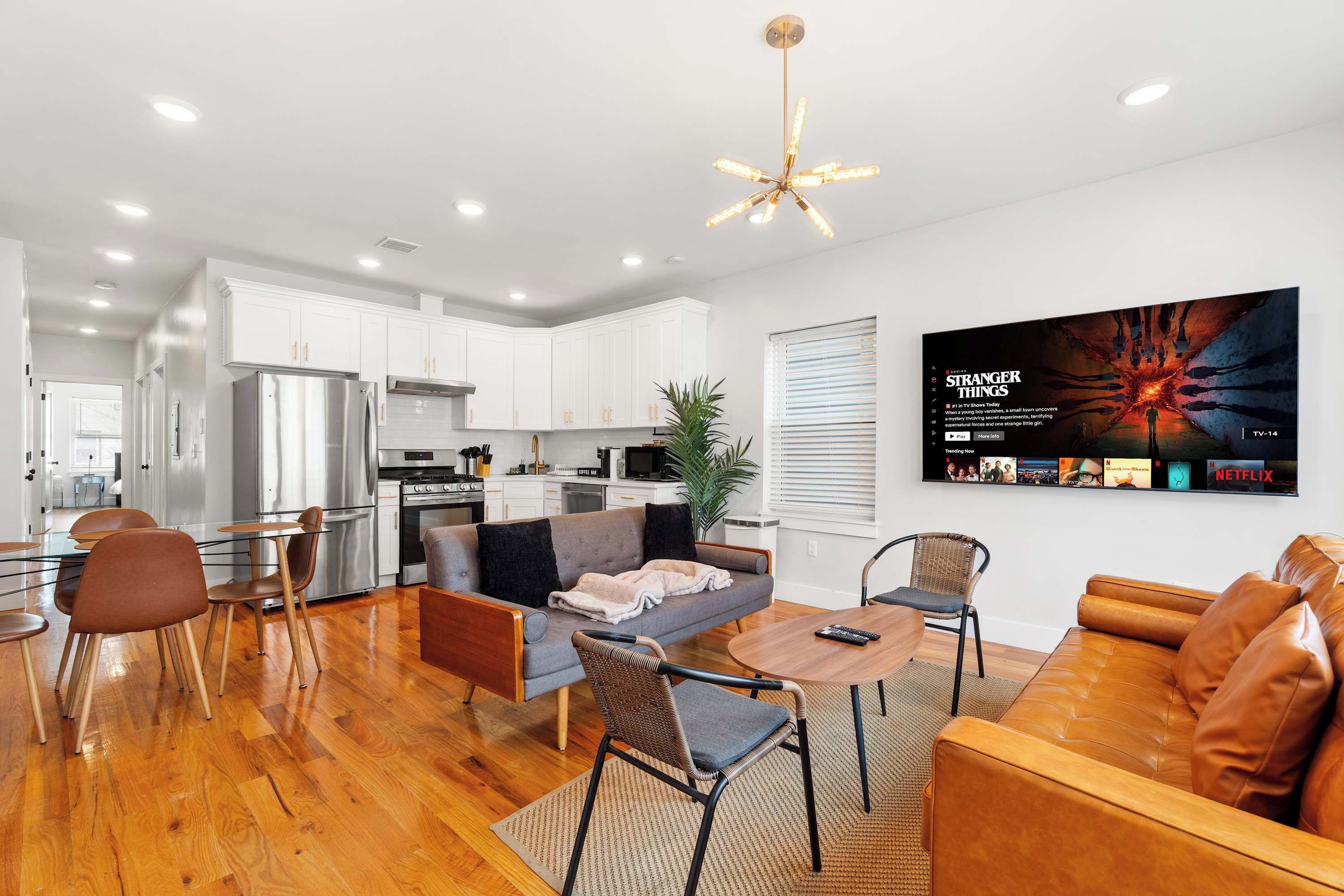 A modern living room with a sofa, coffee table, and chairs, a kitchen with white cabinets, and a TV mounted on the wall displaying a show.