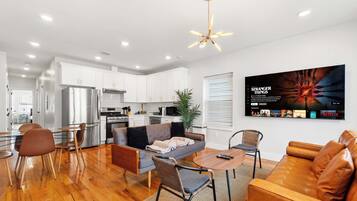 A modern living room with a sofa, coffee table, and chairs, a kitchen with white cabinets, and a TV mounted on the wall displaying a show.