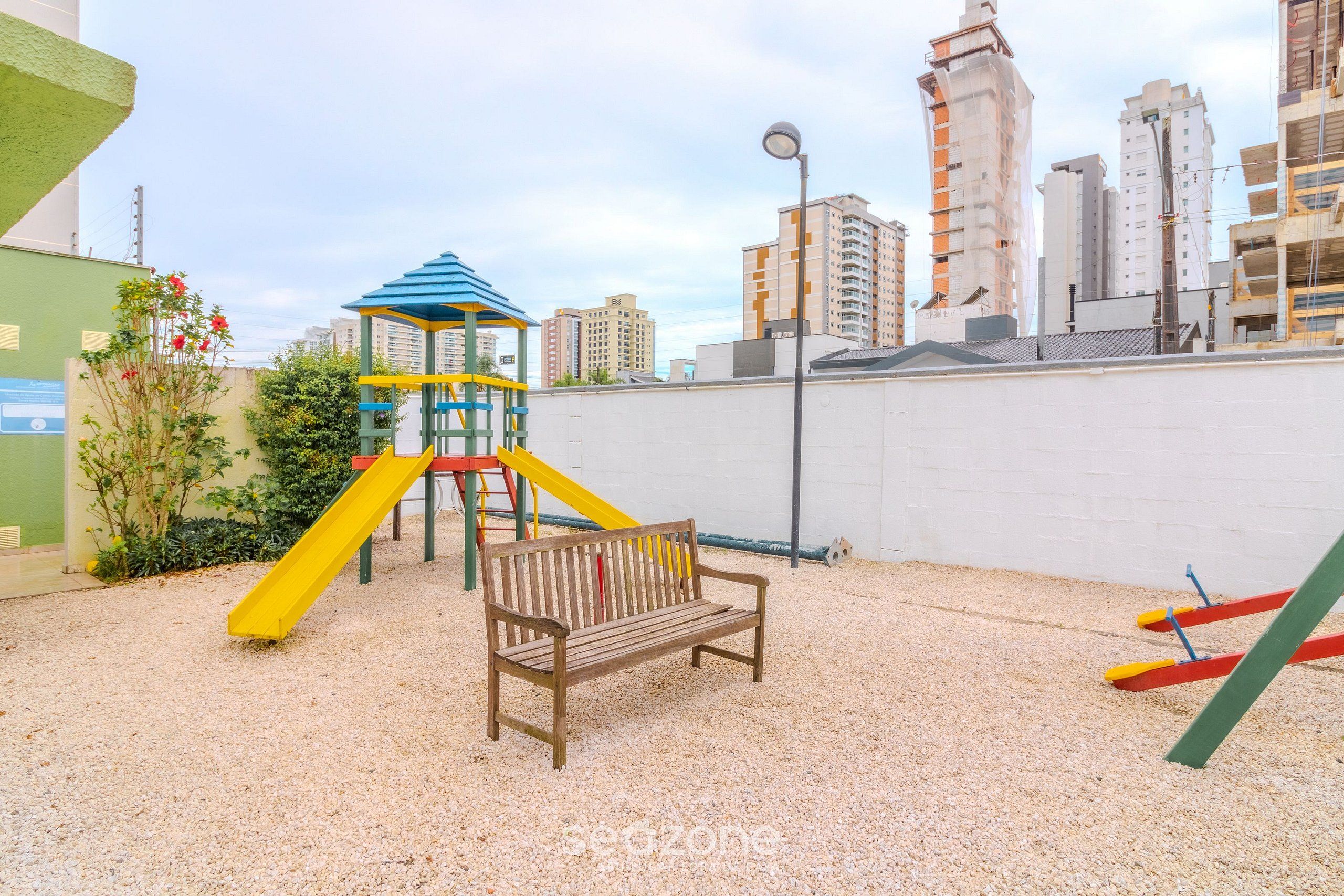 Um parquinho com escorrega e estrutura de escalada, cercado por um muro branco e edifícios ao fundo.