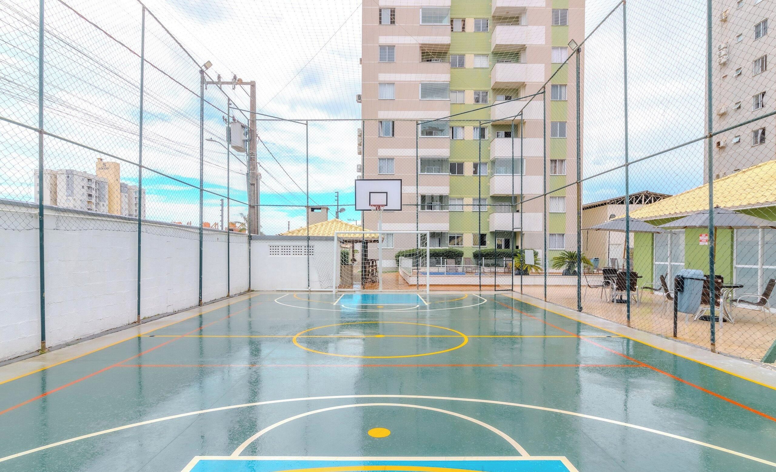 Una cancha de baloncesto cubierta con un aro, rodeada por una cerca de malla metálica, y un edificio de varios pisos al fondo.