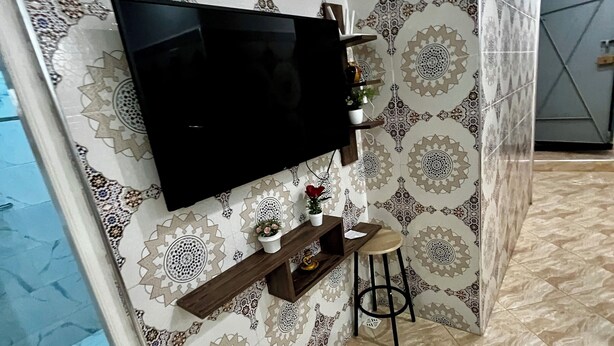 Ein Wohnzimmer mit einem an der Wand befestigten Flachbildfernseher, einem Holzregal mit Dekoration, einem kleinen Hocker und gemusterten Fliesen an den Wänden und auf dem Boden.