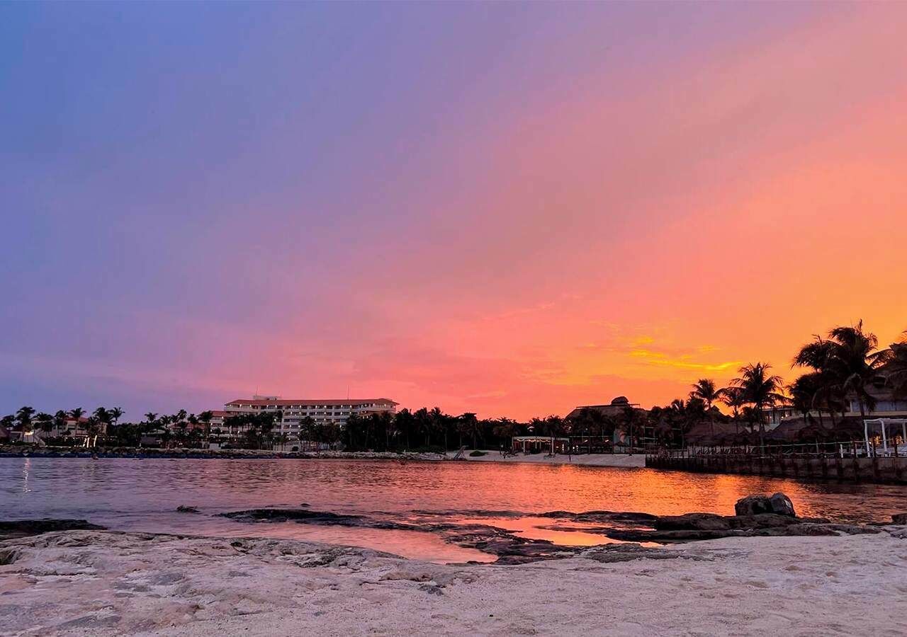 Een strand met een zonsondergang, palmbomen en gebouwen op de achtergrond.