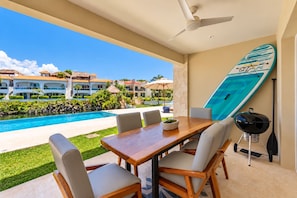 A poolside dining area with a table, chairs, a grill, and a surfboard on the wall. - Lagoonfront · Infinity Pool · Paddleboard (Puerto Aventuras)