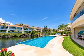 A resort pool area with a clear blue pool, lounge chairs, and a palm tree-lined walkway. - Lagoonfront · Infinity Pool · Paddleboard (Puerto Aventuras)