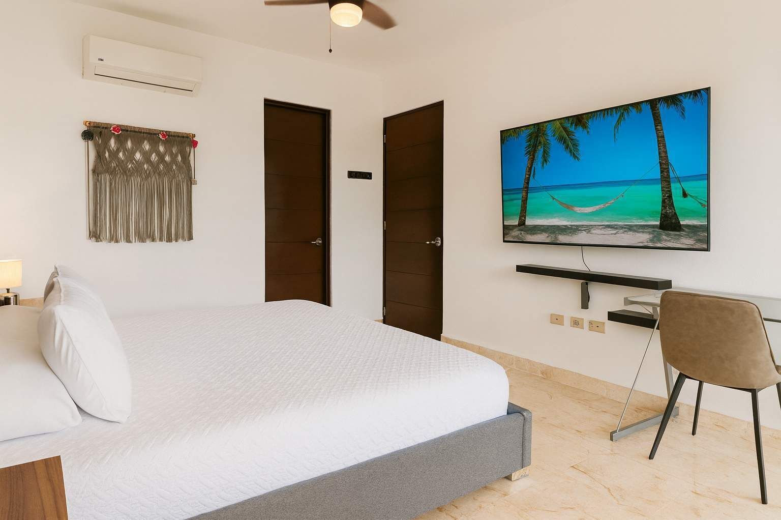 A bedroom with a bed, a desk, a chair, a painting of a beach, and a ceiling fan.