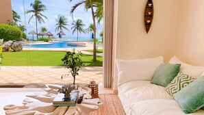 A room with a view of a pool and palm trees, a white sofa with green pillows, and a unique wooden coffee table. - Beachfront|PrivateHotTube|Grill|Groundfloor (Puerto Aventuras)