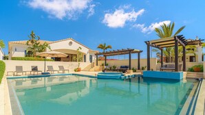 Une piscine dotée de chaises longues et d’une pergola, entourée d’un bâtiment à toit de tuiles et de palmiers.