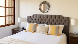 Une chambre à coucher bien aménagée, avec une tête de lit capitonnée, un grand miroir et un lit recouvert de draps blancs et agrémenté de coussins décoratifs.