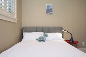 A neatly made bed with white linens, a grey headboard, a small plush toy, a red bedside table with a lamp, and a framed picture on the wall.