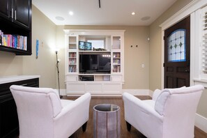 A living room with a white entertainment unit, two white armchairs, a small round table, and a bookshelf.