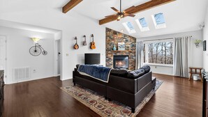 A spacious living room with a stone fireplace, a ceiling fan, and a large window with a view of trees. - One-level w/FirePit, 2 Living Areas & Resort Amens (Hedgesville)