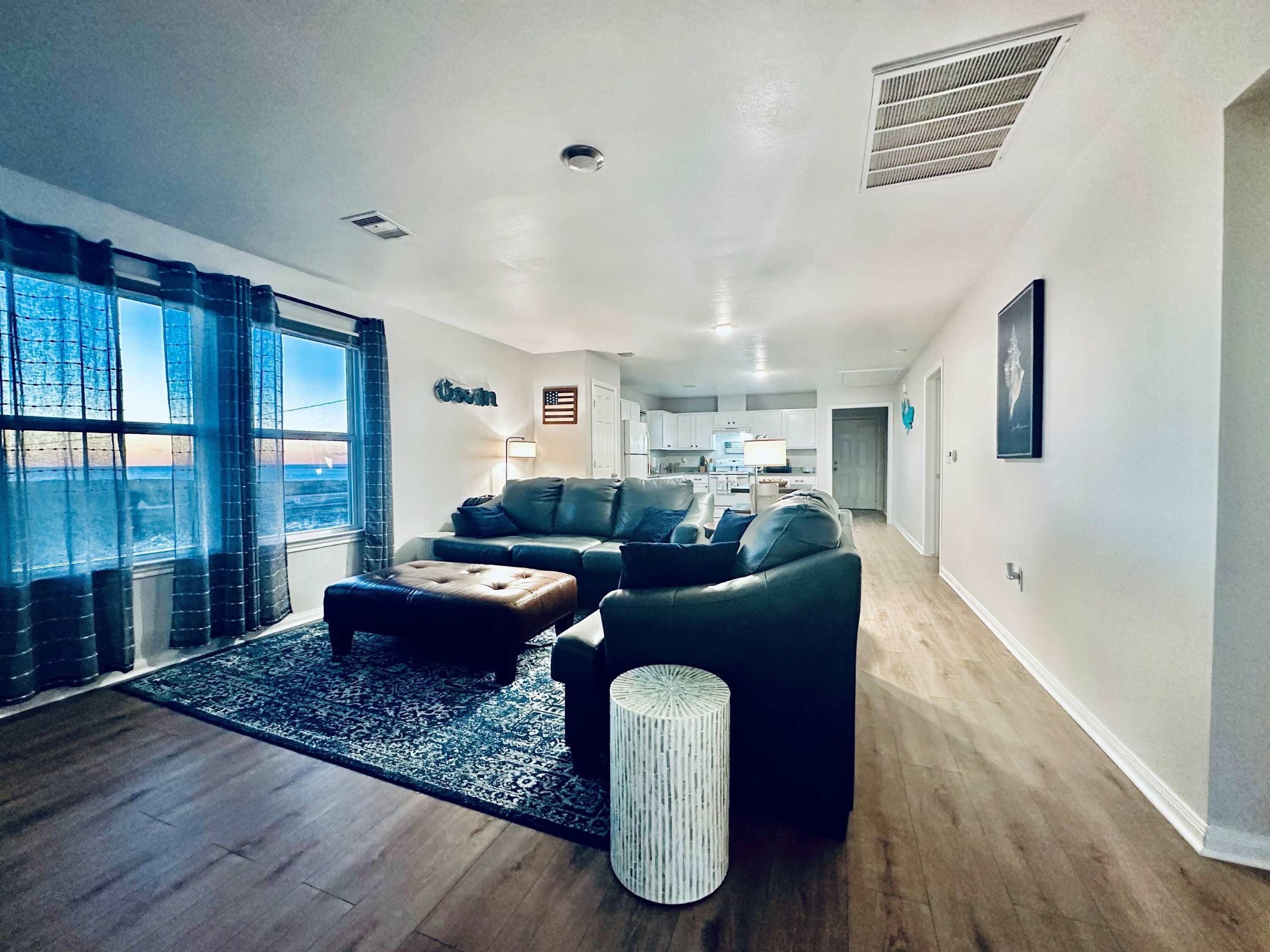 A modern living room with a sofa, ottoman, and a small side table. There is a window with curtains, a rug, and a door leading to a kitchen.