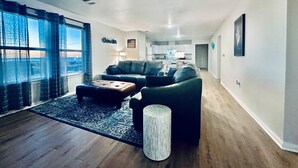 Ein modernes Wohnzimmer mit einer Couch, einem Hocker und einem kleinen Beistelltisch. Ein Fenster mit Vorhängen, ein Teppich und eine Tür, die in eine Küche führt.