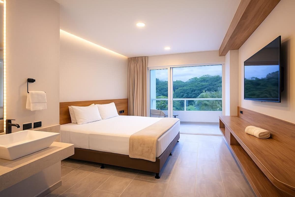 Une chambre d’hôtel moderne dotée d’un grand lit, d’une télévision intégrée, d’une salle de bain avec un lavabo et un miroir, et d’un balcon offrant une vue sur la verdure.