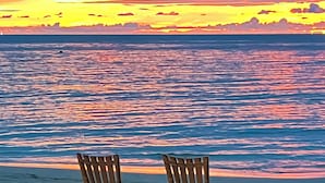 Deux chaises en bois, placées face à une plage, avec un coucher de soleil sur l’océan.