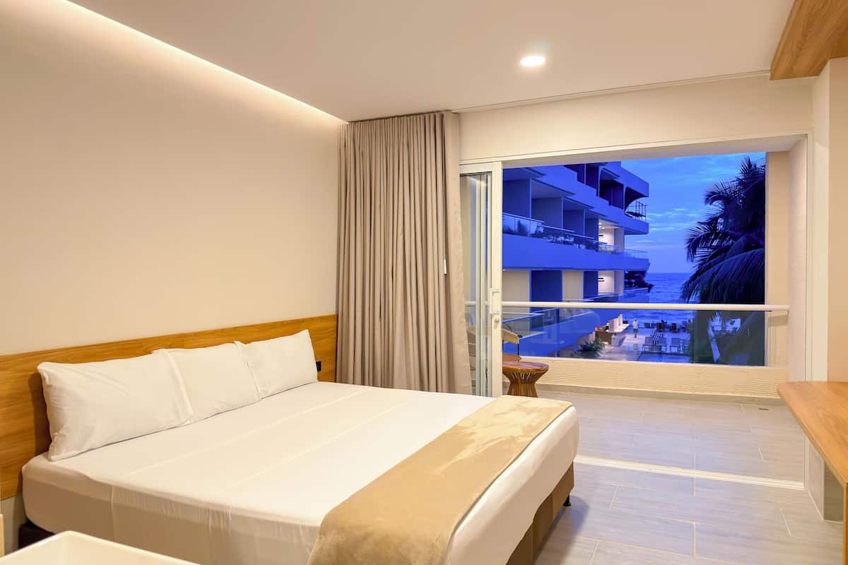 Une chambre d’hôtel avec un lit, une petite table et un balcon donnant sur la piscine et les bâtiments environnants.