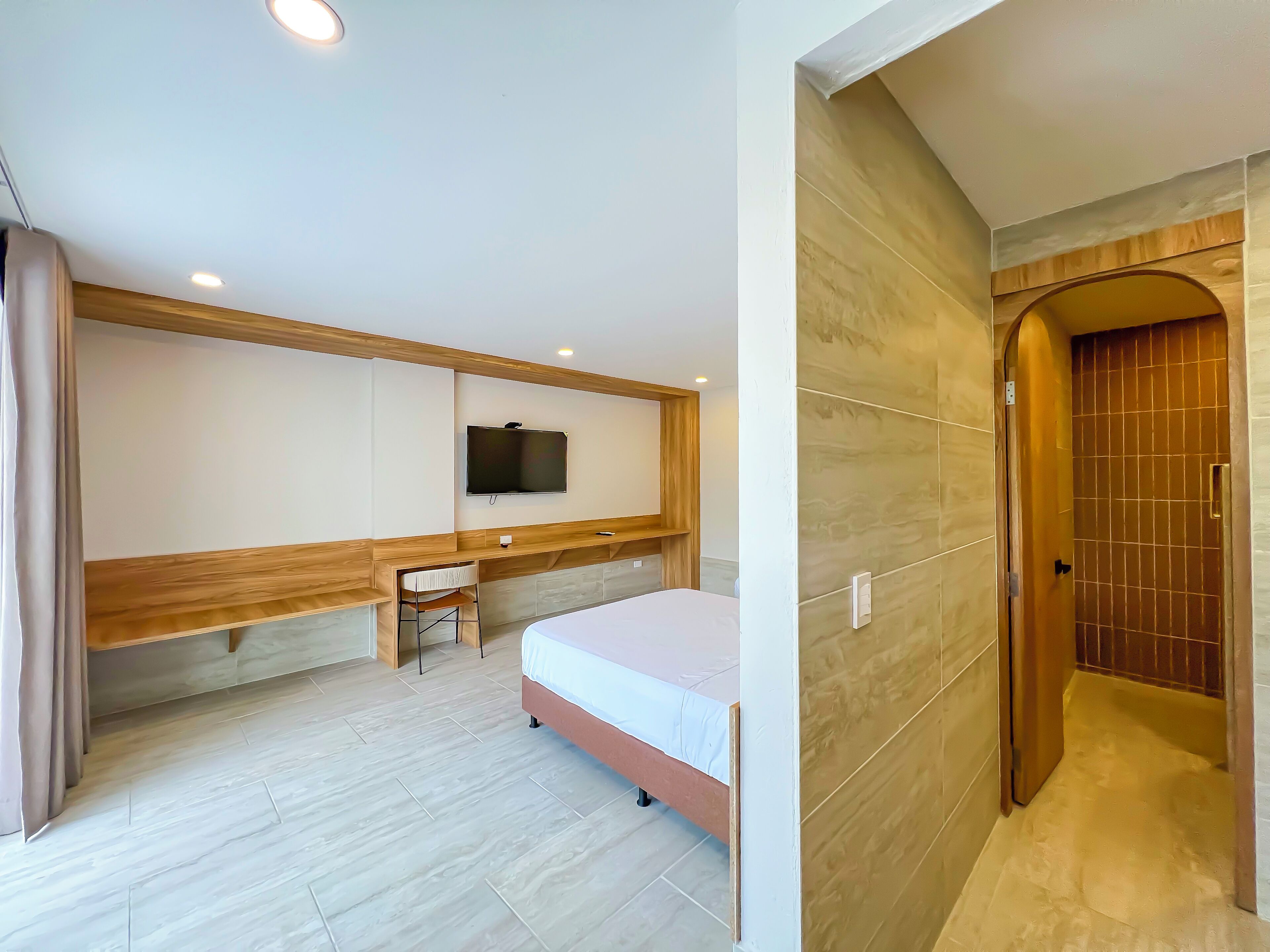 A modern hotel room with a bed, a desk, a chair, a TV mounted on the wall, and a wooden door.