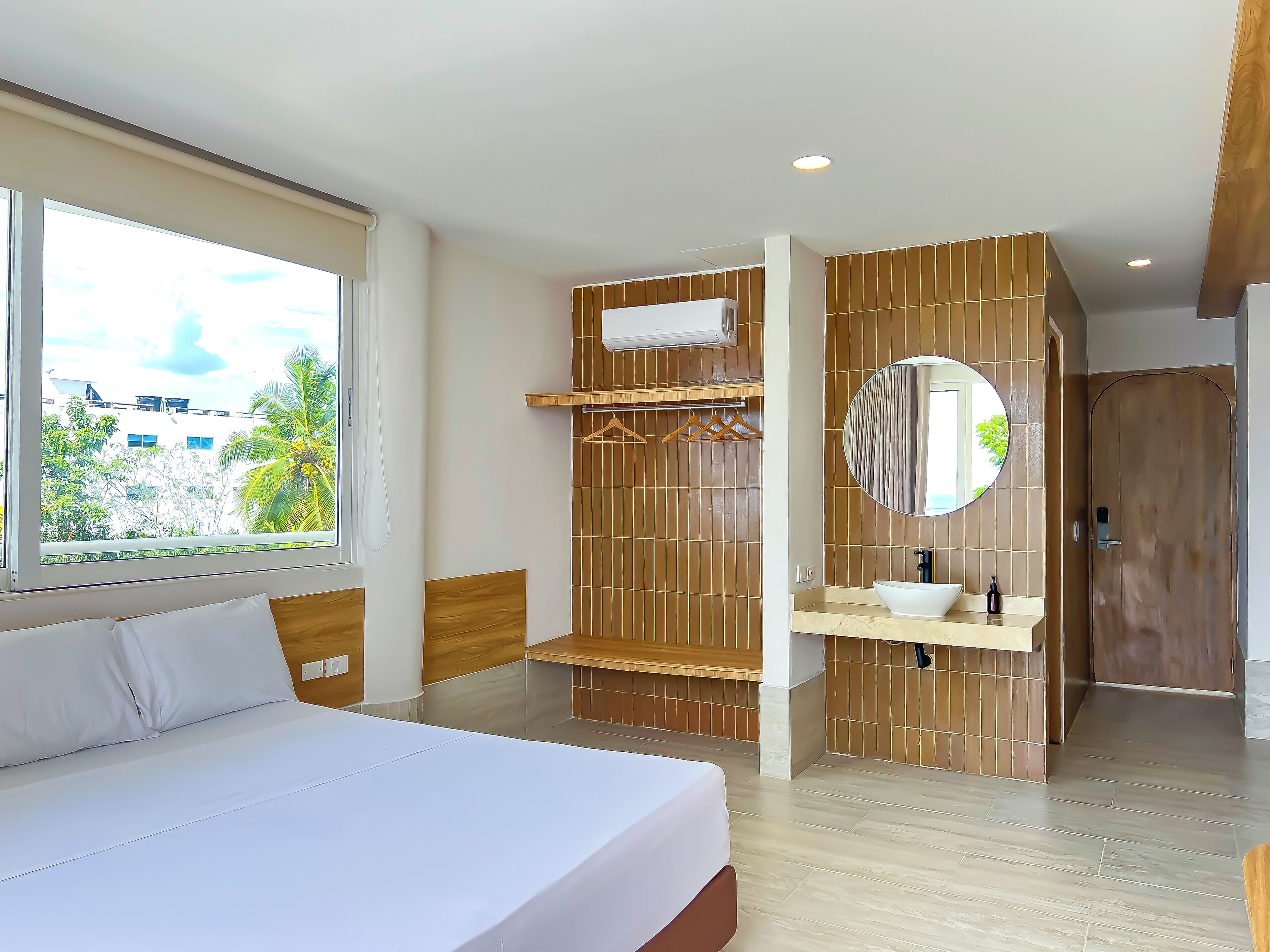 A modern hotel room with a large bed, a bathroom with a wooden vanity, a round mirror, and a window with a view of buildings and trees.