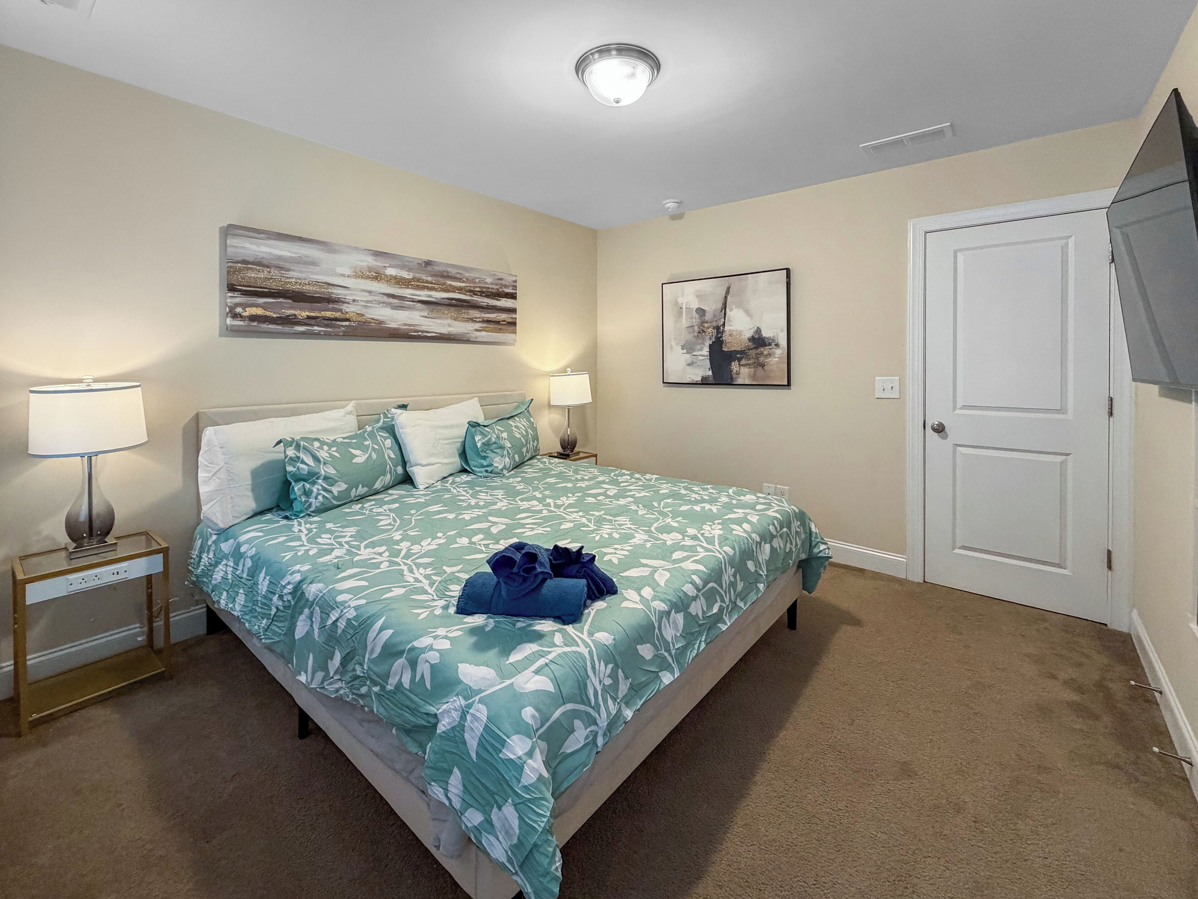 A bedroom with a bed, two bedside tables with lamps, a door, and two wall-mounted artworks.