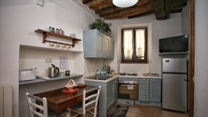 A compact kitchen with a wooden dining table, white chairs, a built-in oven, a refrigerator, and a window with curtains. - Il Terrazzino (Cetona)