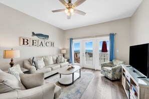A living room with a sofa, armchairs, a coffee table, a TV, and a ceiling fan. - Beachfront Duplex | Heated Pool | Sleeps 27 (Gulf Shores)