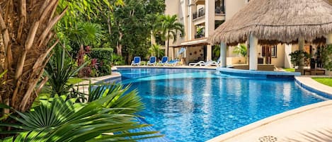 A swimming pool with lounge chairs, surrounded by tropical plants and a building with balconies.
