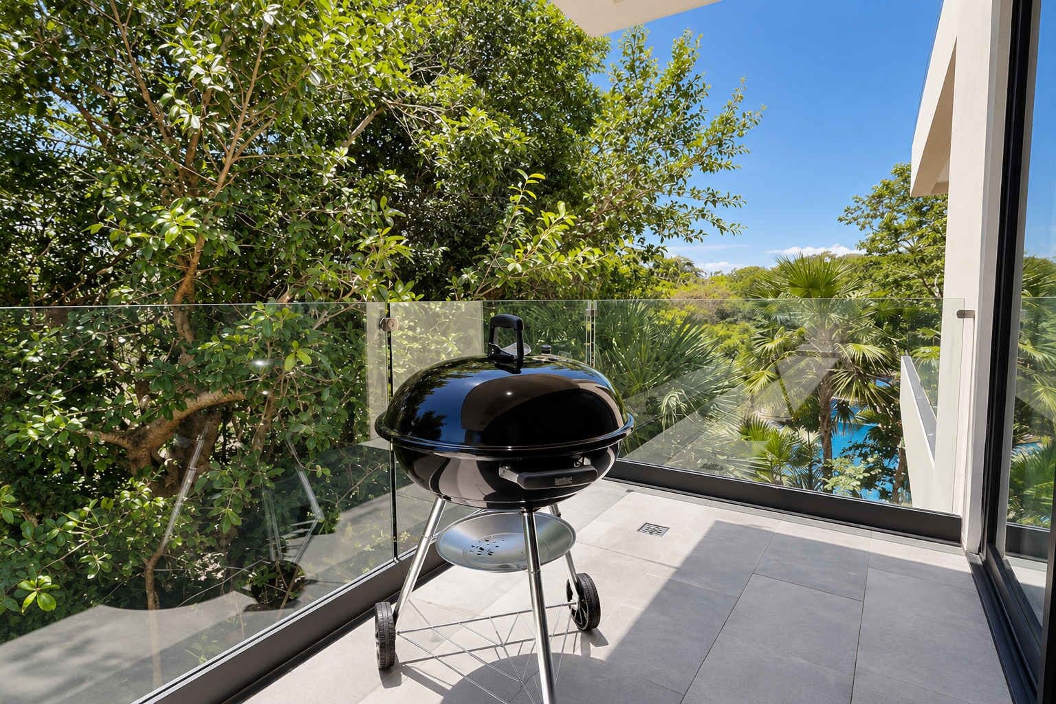 A balcony with a barbecue grill, glass railing, and a view of a lush garden and pool.
