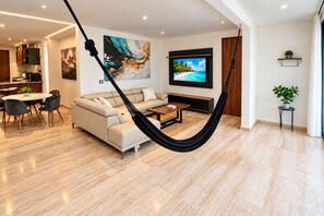 Una sala de estar moderna con sofá, mesa de centro y una hamaca. Hay un televisor montado en la pared y una zona de comedor con sillas.