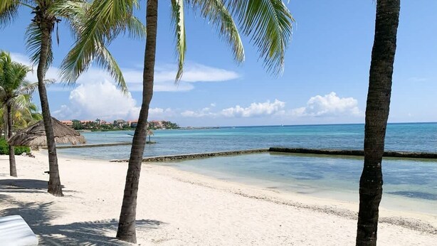 A beach with white sand, palm trees, and a clear blue sea. - Ground Floor Condo with Marina View & Pool Access (Puerto Aventuras)