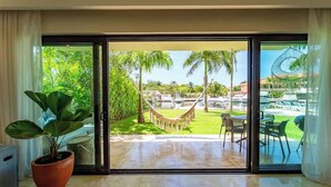 A room with a large sliding glass door leading to a patio with a hammock, palm trees, and a pool area. - Ground Floor Condo with Marina View & Pool Access (Puerto Aventuras)
