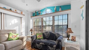 Een woonkamer met een bank, een fauteuil, een kleine tafel en een raam met jaloezieën.