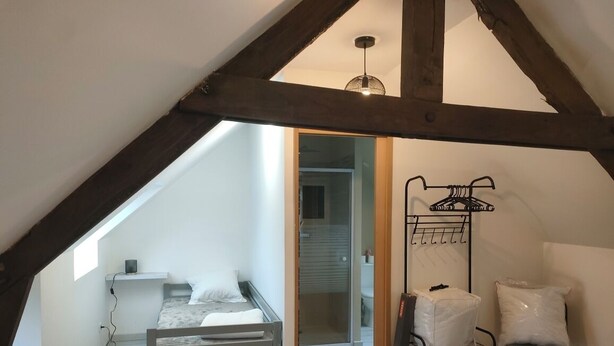 Un dormitorio con techo inclinado, una cama, una silla y un perchero metálico con almohadas.
