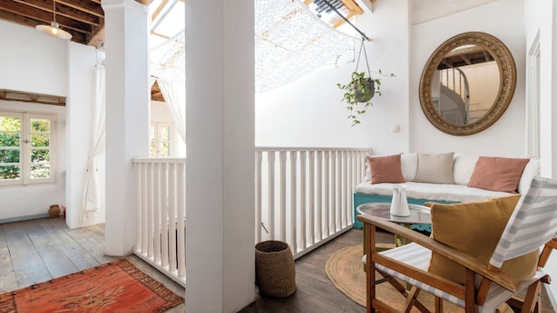 Una habitación acogedora con techo blanco, vigas de madera, barandilla blanca, un sofá con cojines, una planta colgante y un espejo redondo en la pared.
