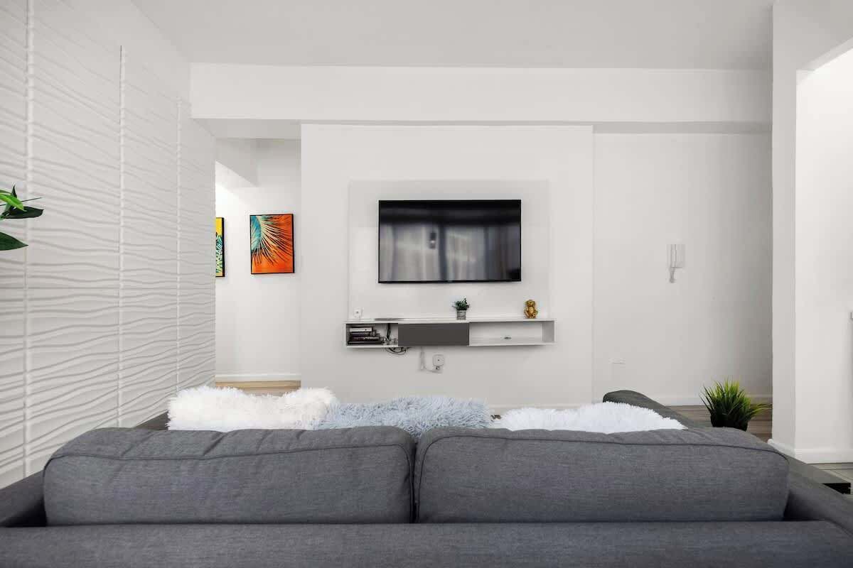 A modern living room with a grey sofa, a mounted TV, a white wall, and a small shelf with decorative items.