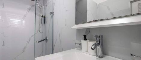 Un baño moderno con pared de mármol blanco, ducha envuelta en vidrio y lavabo blanco con espejo superior.
