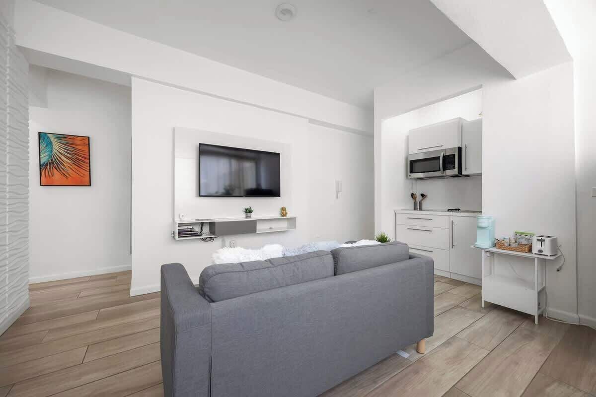 A modern living room with a grey sofa, a mounted TV, and a kitchen area with white cabinets and a microwave.