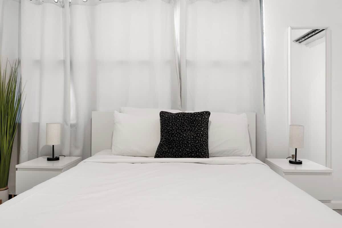 A neatly made bed with white linens and a black patterned pillow, flanked by white nightstands with lamps, in a room with sheer white curtains and a mirror on the wall.