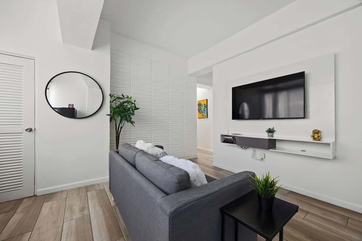 A modern living room with a grey sofa, a round mirror, a flat-screen TV, and a small black table with a plant.